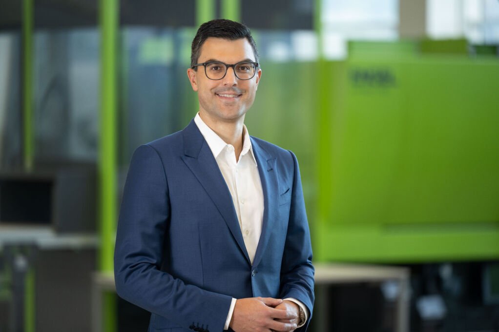 Stefan Engleder, CEO of the ENGEL Group, smiles in a blue suit, with a green injection moulding machine out of focus behind him.